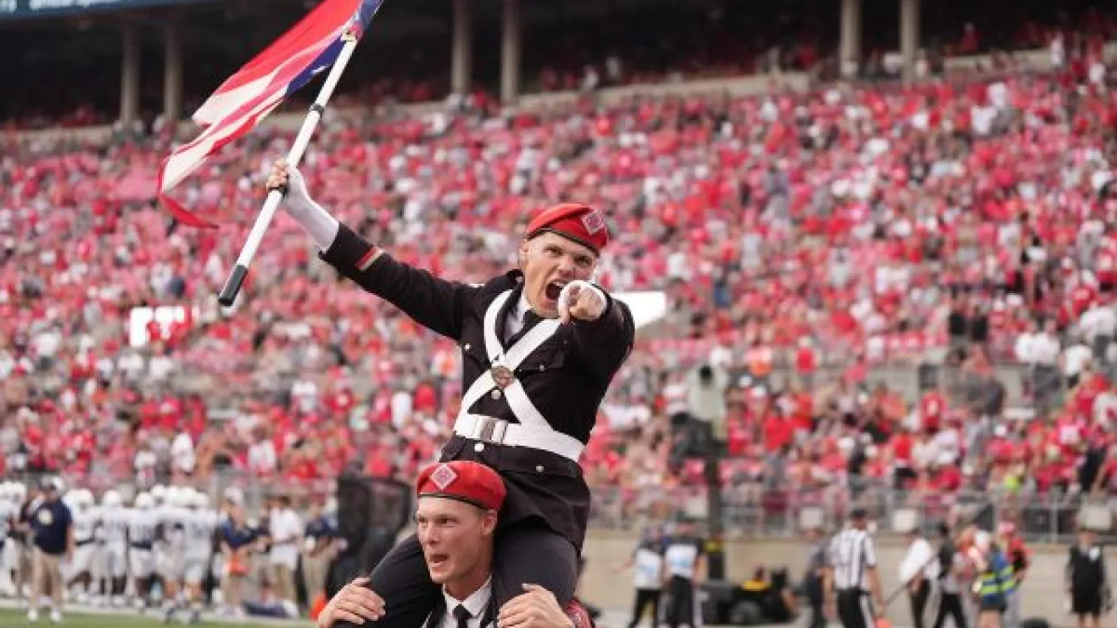 a young man in a marching band uniform sits on the shoulders of a drummer, his right hand grips an Ohio flag, his left points in the direction of the touchdown