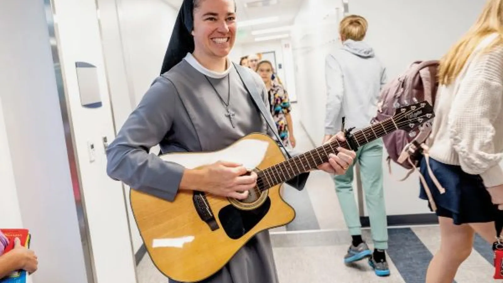 Sister M. Xavier Schulze smiles while playing guitar in a school hallway