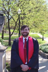 Owen White in graduation regalia standing in front of greenery.