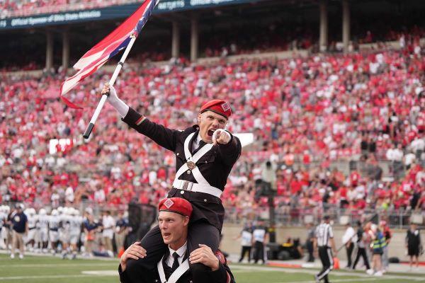 a young man in a marching band uniform sits on the shoulders of a drummer, his right hand grips an Ohio flag, his left points in the direction of the touchdown
